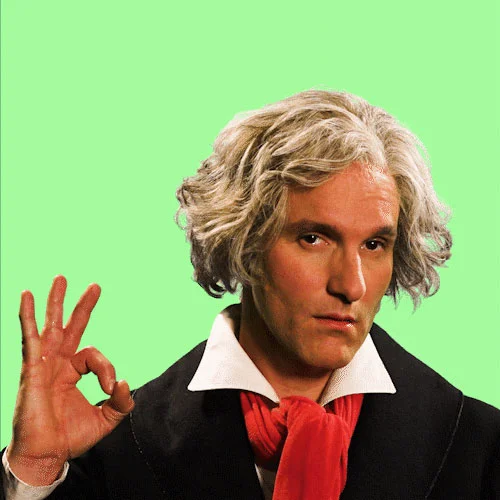 Ein Mann mit längeren grauen Haaren steht vor grünem Hintergrund und formt seinen Daumen und Zeigefinger zu einem Kreis.